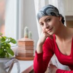 Young female cancer patient wearing a head scarf, sitting contemplatively in front of window.