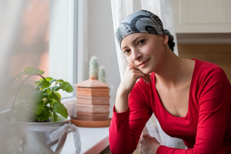 Young female cancer patient wearing a head scarf, sitting contemplatively in front of window.
